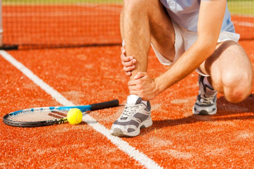 Man on tennis court kneels down holding his ankle