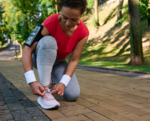 Active middle-aged woman runner, African American athlete tying shoelaces getting ready for jog along the forest park on a warm sunny summer day.