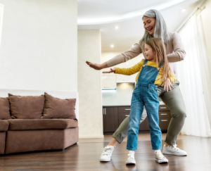 Joyful grandmother dancing and smiling while having fun together with her little granddaughter in a living room at home.