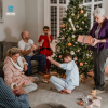 Three generation family are opening presents together on Christmas morning. They are sitting by the Christmas tree in the living room of their home, wearing pajamas.